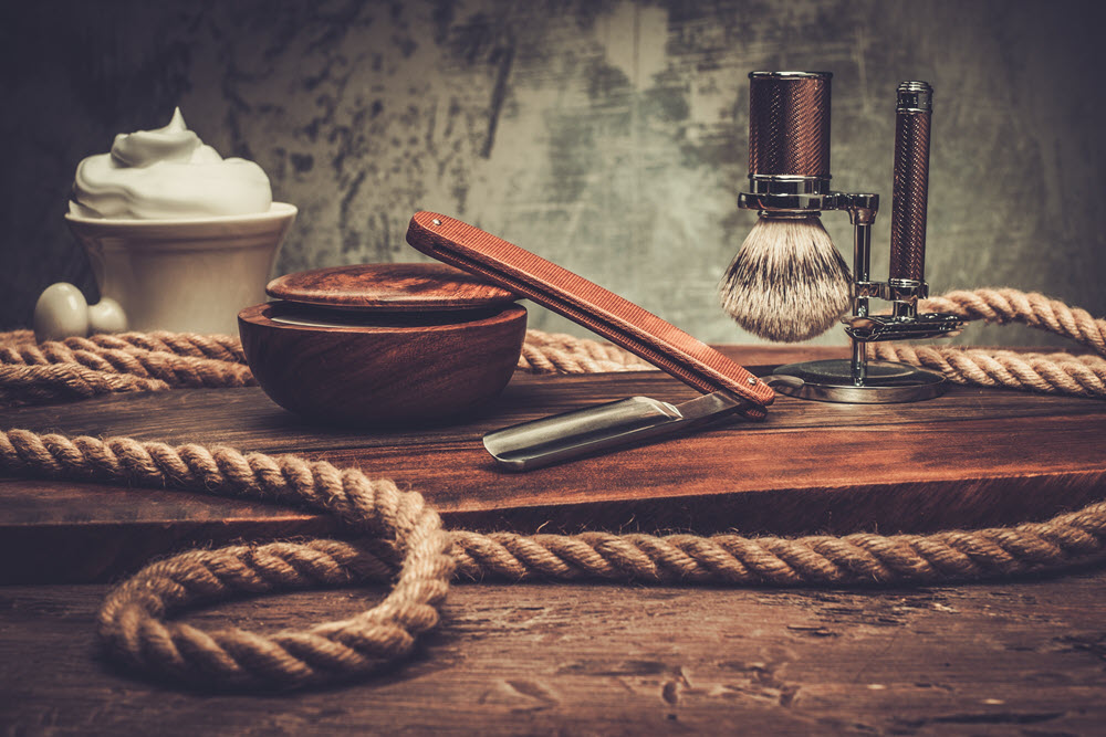 Picture of a wooden shaving bowl, a cermaic shaving bowl with lathered cream, a straight razor, a stand with a shaving brush and a safety razor on a wooden surface. A rope is laid around the items.