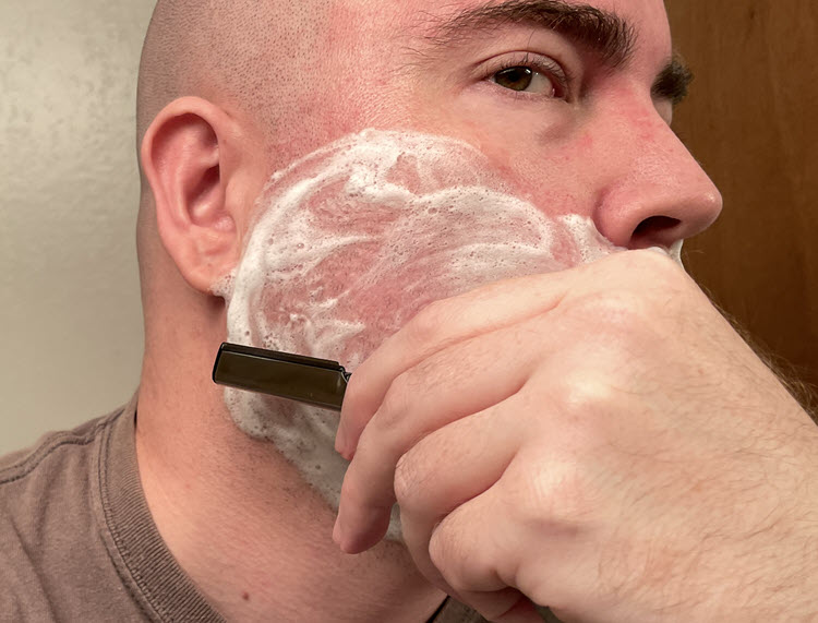 A man about to shave the right side of his lathered face with a Dovo Shavette with a black insert and Dovo long blades going against the grain.