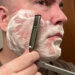 A man with a lathered face about to shave across the grain on his right cheek with a Dovo Shavette