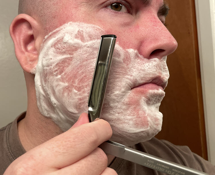 A man with a lathered face about to shave across the grain on his right cheek with a Dovo Shavette