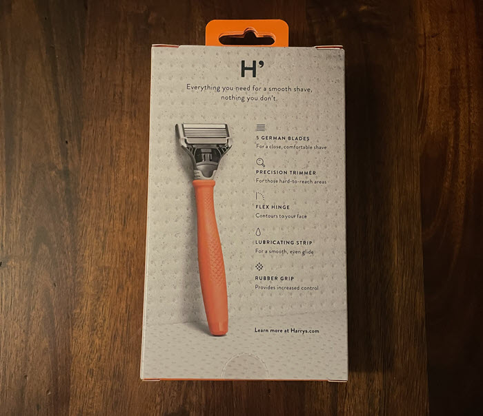 Back of Harry's razor packaging stating "Everything you need fo a smooth shave, nothing you don't." An image of the bright orange model razor on the left hand side. Key selling points are listed on the right side which are "5 German blades For a close, comfortable shave", "Precision Trimmer for those hard-to-reach areas", "Flex hinge contours to your face", "Lubricating strip for a smooth, even glide", and "Rubber Grip provides increased control".