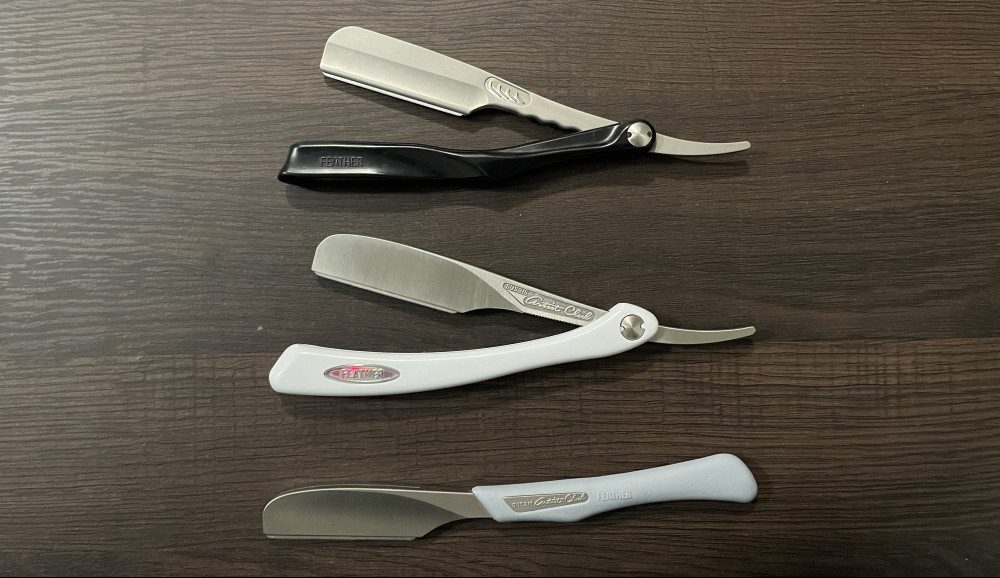 Three Feather Artist Club Razors on a dark wood background. A folding black-handled SS razor, folding DX razor and non-folding DX razor